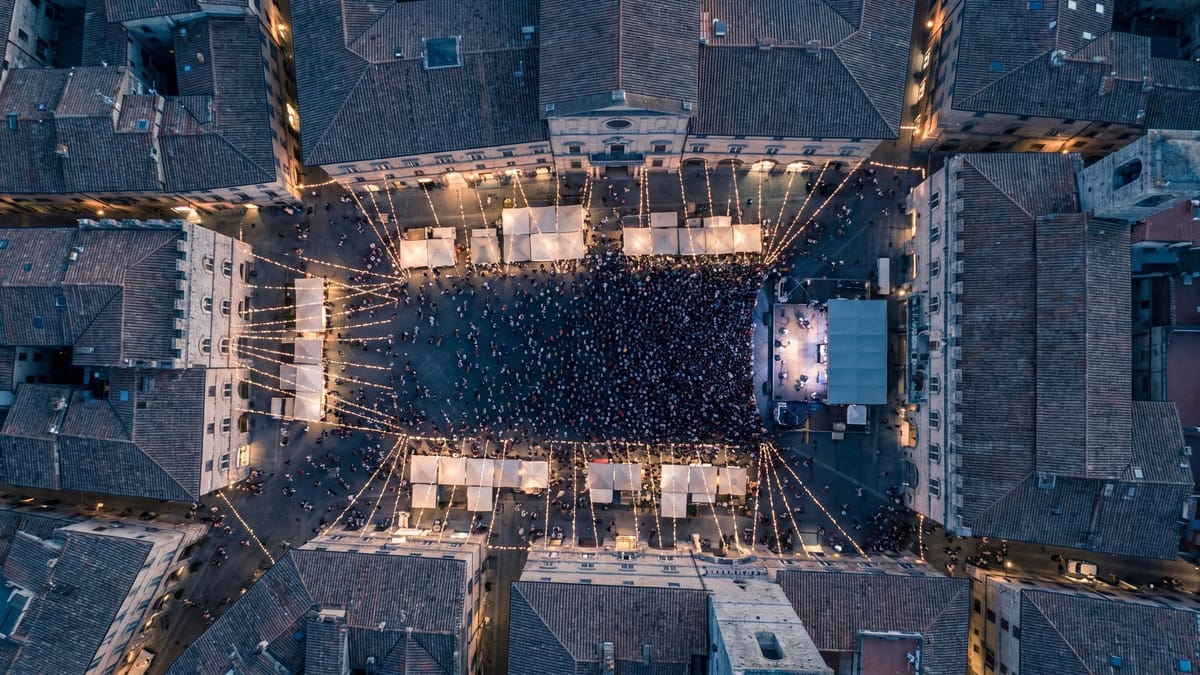 Veduta aerea dall'alto di una piazza italiana durante una festa, con bancarelle, palco e folla indistinta